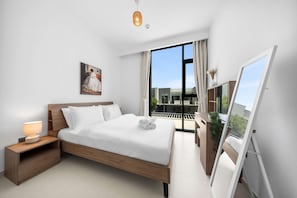 Ein modernes Schlafzimmer mit einem großen Bett, einem Nachttisch mit Lampe, einem Gemälde an der Wand und Blick ins Freie durch ein großes Fenster.