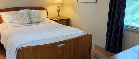 A bedroom with a wooden bed, a bedside table with a lamp, a framed picture on the wall, and a window with curtains.