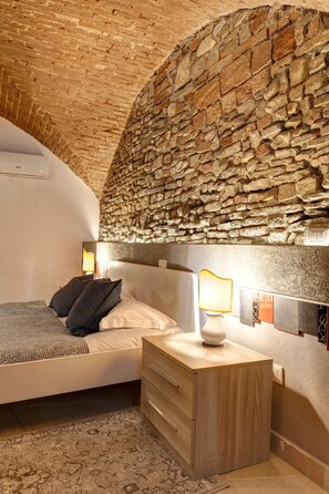 Ein Schlafzimmer mit Steinwand, einem Bett mit Kissen, einem Nachttisch und einem Beistelltischchen mit einer Lampe.