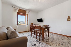 Ein Wohnzimmer mit einer Couch, einem Esstisch mit Stühlen, einem an der Wand montierten Fernseher und einem Fenster mit Vorhängen.