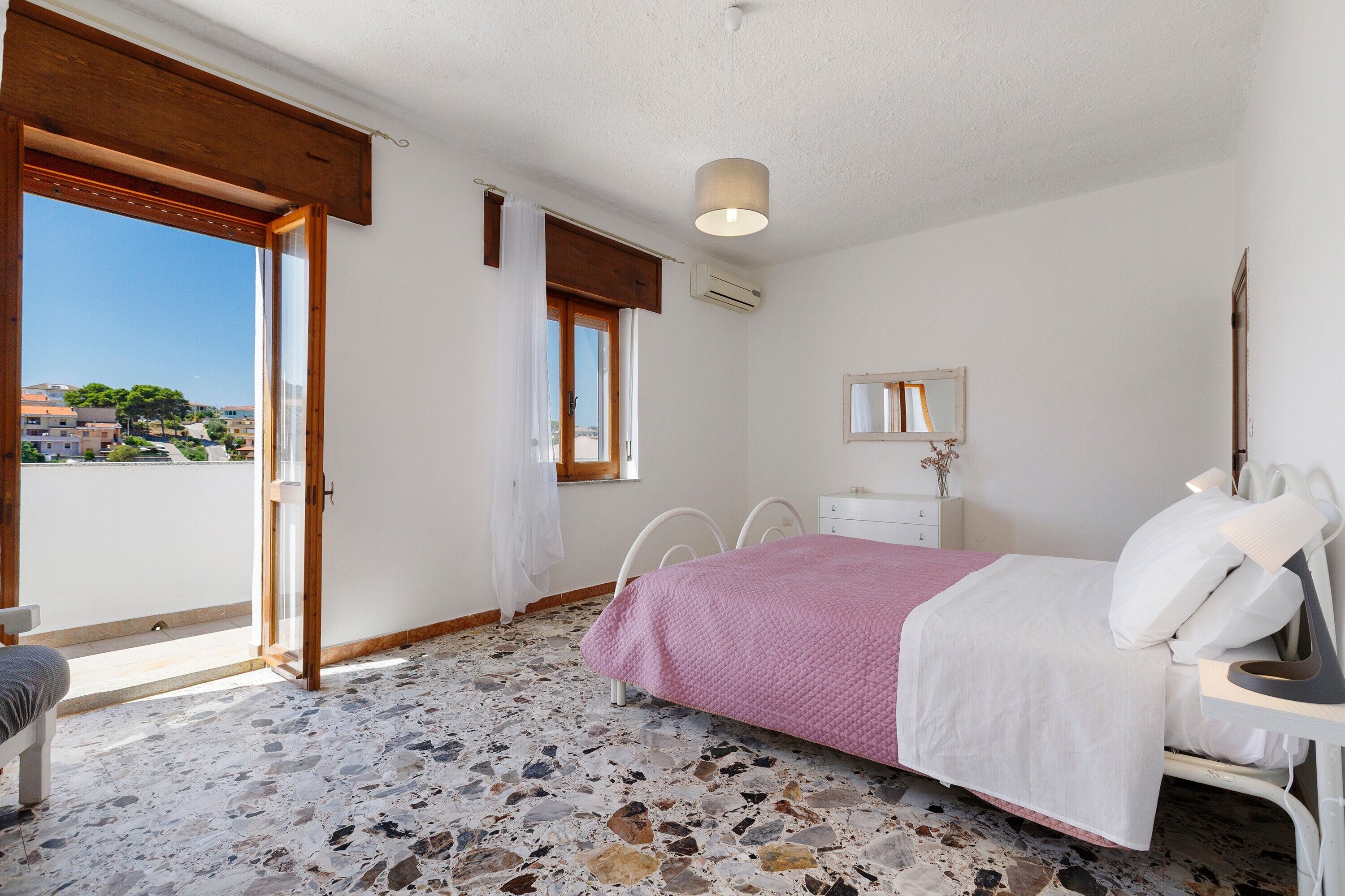 Une chambre à coucher avec un lit, une commode, un miroir et une fenêtre donnant sur des immeubles.