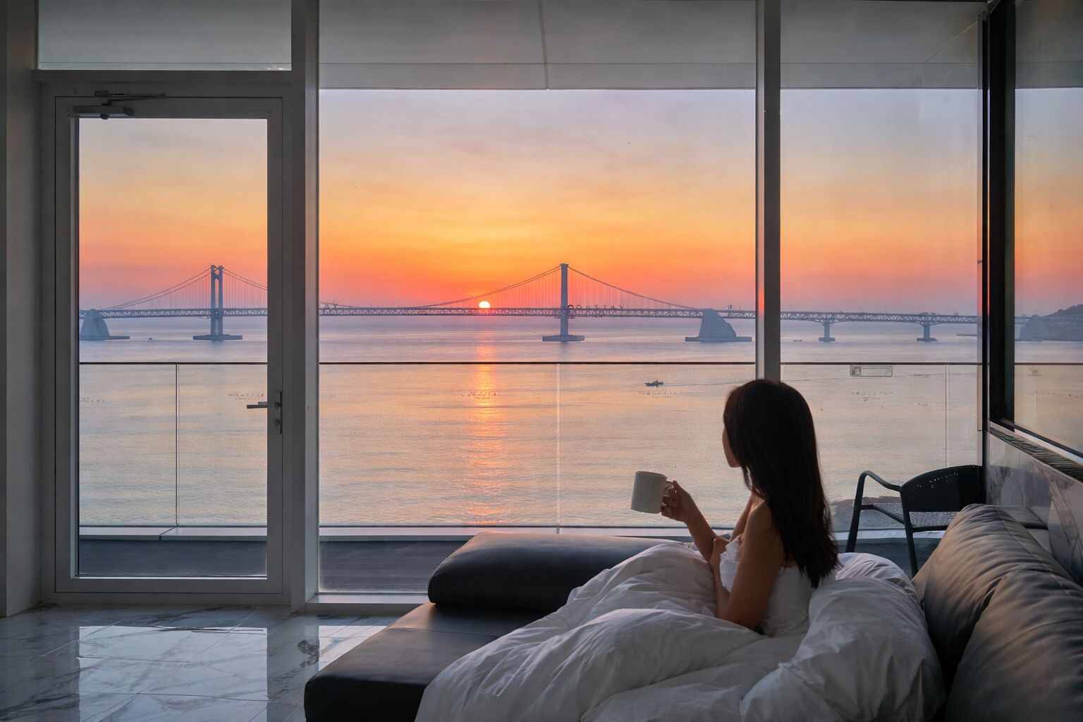 Una habitación con un ventanal grande que da a un atardecer con un puente, una cama con almohadas y una persona leyendo.