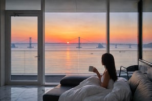 A room with a large window overlooking a sunset with a bridge, a bed with pillows, and a person reading.