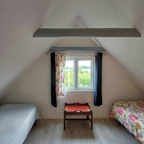 Ein kleines, einzelnes Zimmer mit schrägen Wänden, einem Fenster mit Vorhängen, einer hölzernen Bank und einem Bett mit Blumenmuster-Decke.