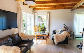 A cozy living room with a leather sofa, a coffee table with books, a guitar, and a view of greenery through the windows. - Cottage in the garden of Fejø Organic Food Forest (Fejø)