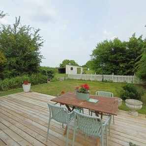 Eine Holzterrasse mit Tisch und Stühlen, umgeben von viel Grün und einem weißen Zierzaun.