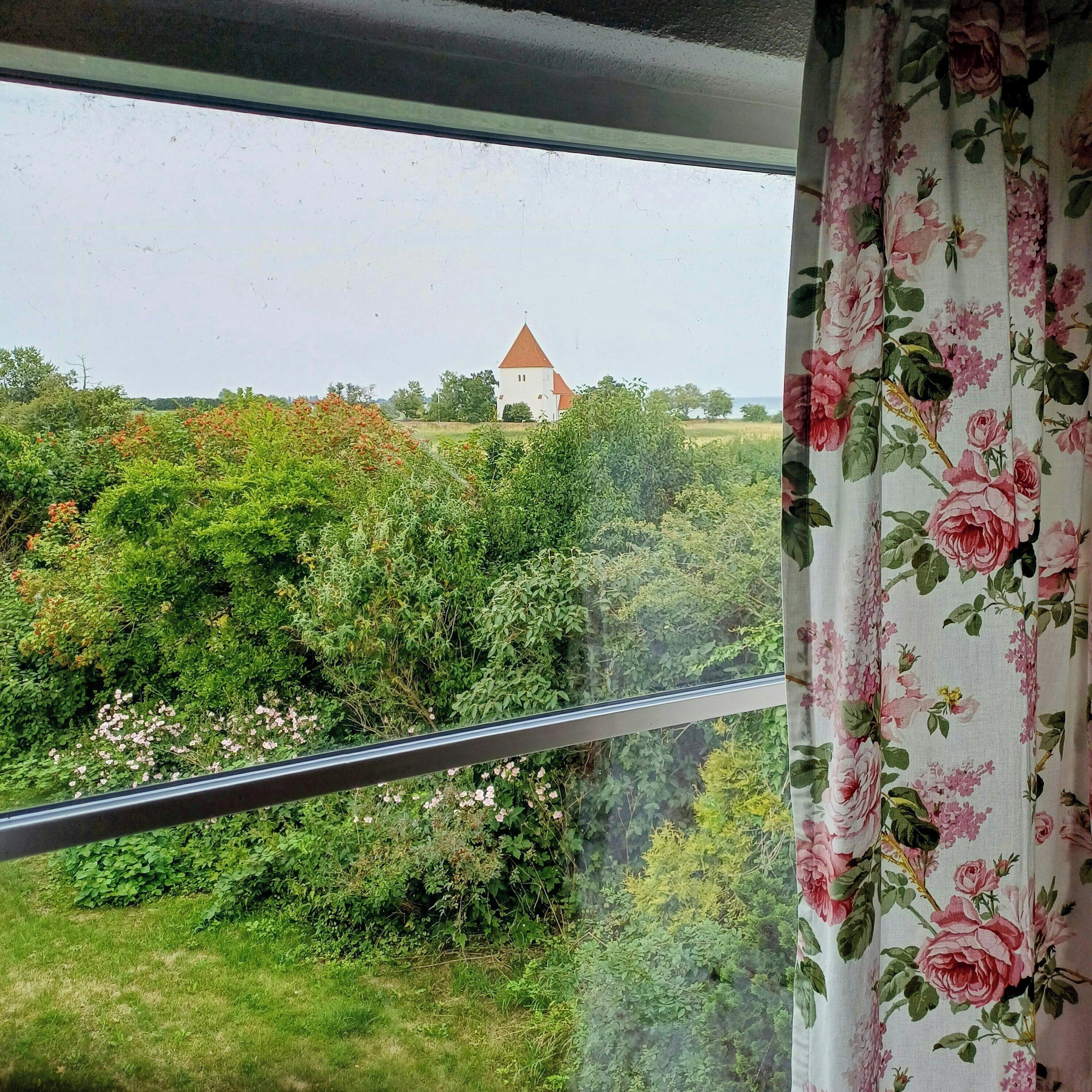 Uitzicht door een raam met een bloemen gordijn, waarop een gebouw met rood dak en een toren te zien is, omringd door weelderige begroeiing.