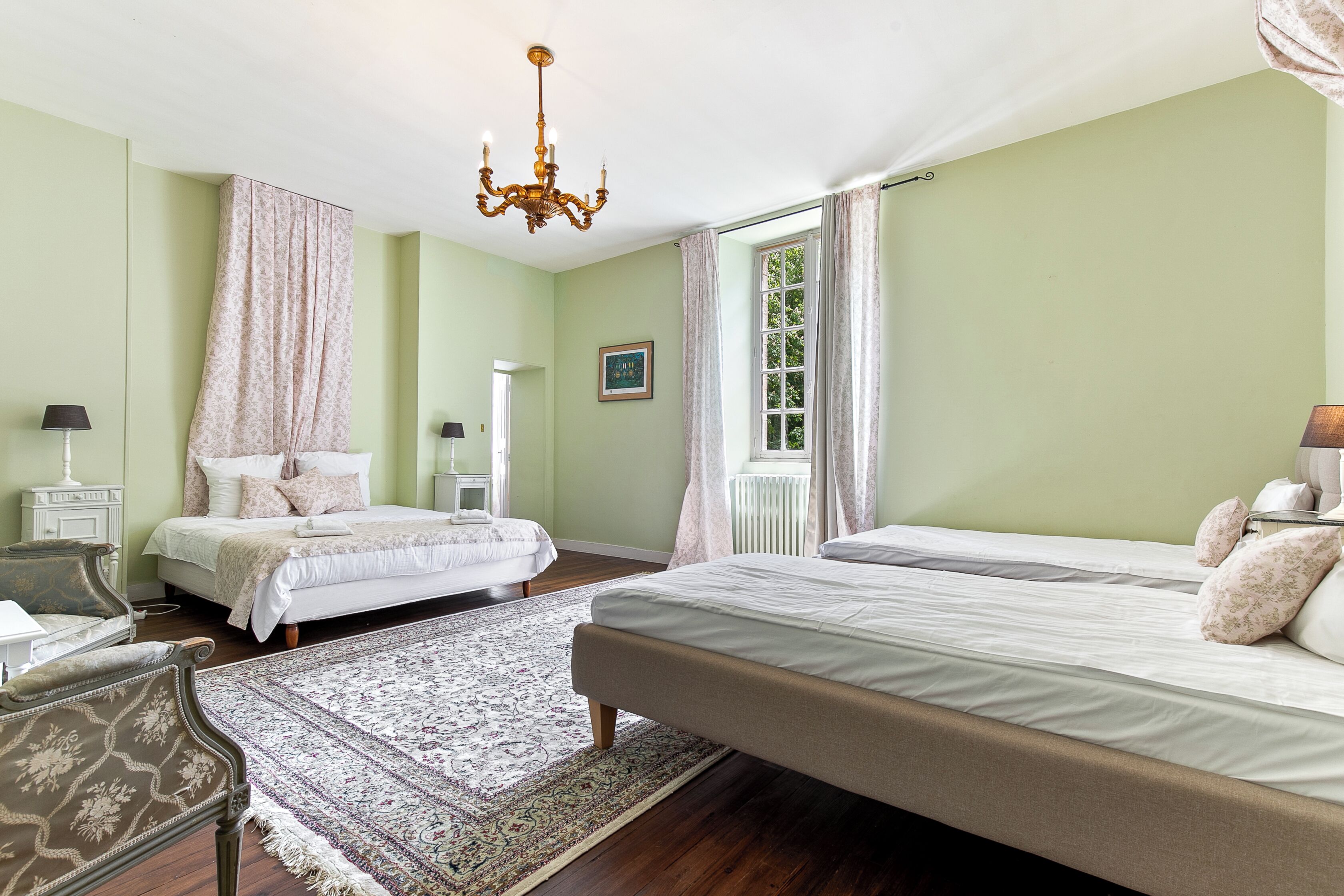 A bedroom with two beds, a chandelier, a window with curtains, a bedside table, and a chair.