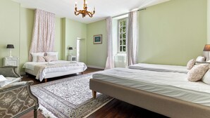 Quarto com duas camas, um candelabro, uma janela com cortinas, uma mesinha de cabeceira e uma cadeira.