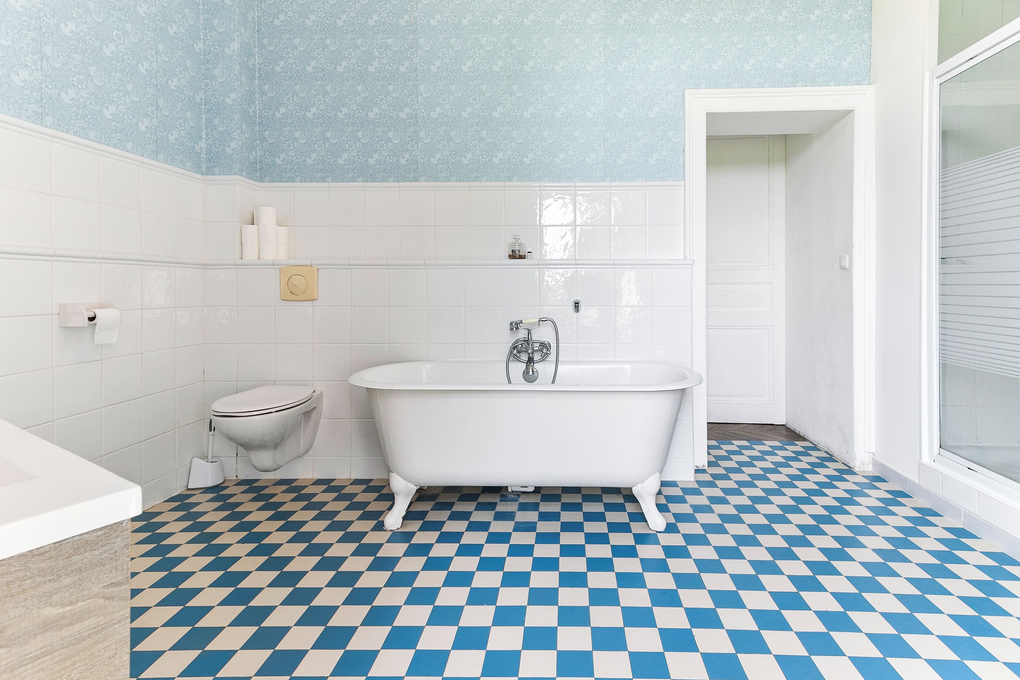 Un bagno con pavimento a scacchi, una vasca da bagno bianca, un water e una parete con motivo blu.