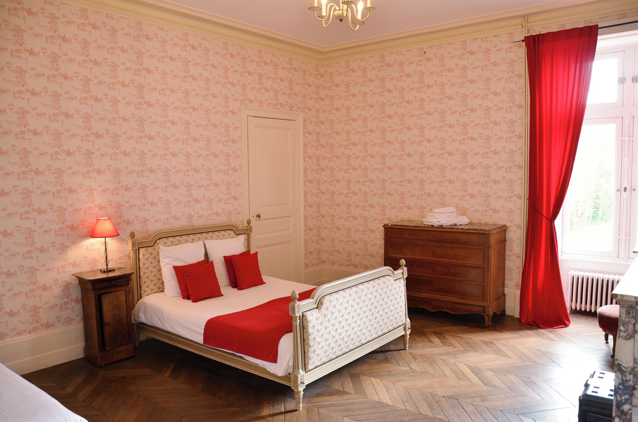 Ein Schlafzimmer mit Bett, Kommode, Nachttisch und einem Fenster mit roten Vorhängen.