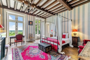 Ein Schlafzimmer mit einem Himmelbett, einer Wand im Rot-Weiß-Streifenmuster, einem großen Fenster und Blick ins Freie.