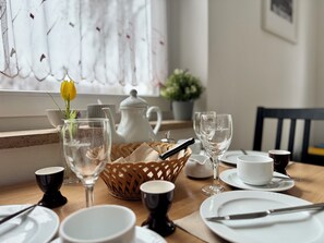 A dining table set with white plates, bowls, and glasses, a white teapot, a wicker basket, and a yellow tulip in a glass vase.