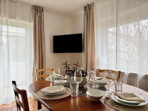 Un coin repas avec une table en bois dressée pour quatre personnes, un téléviseur à écran plat fixé au mur et de grandes fenêtres ornées de rideaux légers.