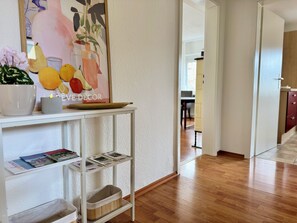 Un couloir avec un mur blanc, un plancher en bois, une table blanche avec un présentoir à magazines, une plante en pot et une œuvre d’art encadrée.