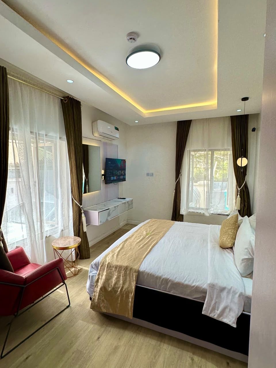 Une chambre d’hôtel avec un lit, une chaise rouge, une petite table, un téléviseur et une fenêtre avec des rideaux.