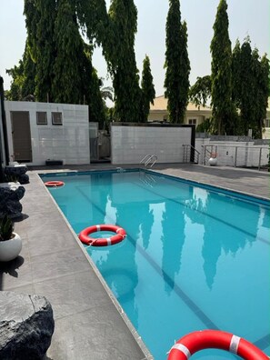 Piscina com boias salva-vidas, rodeada por um deck de azulejos e árvores altas.
