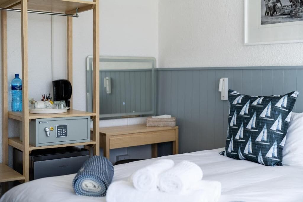 A hotel room with a bed, a bedside table, a safe, a mirror, a towel rack, and a wall-mounted picture.
