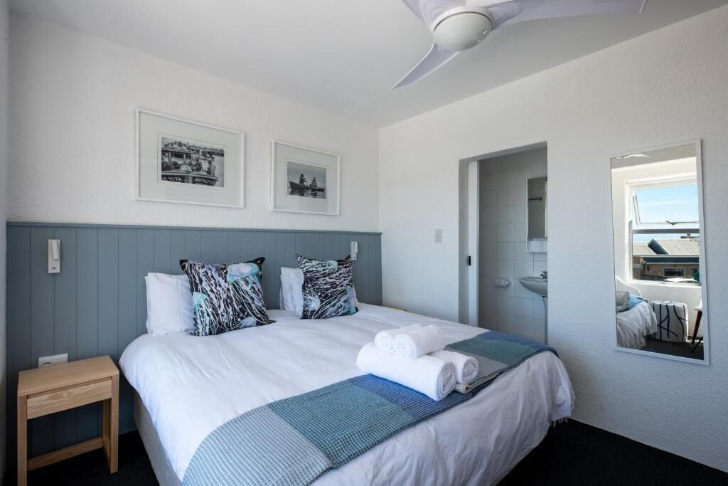 A hotel room with a bed, a small table, a ceiling fan, and two framed pictures on the wall.