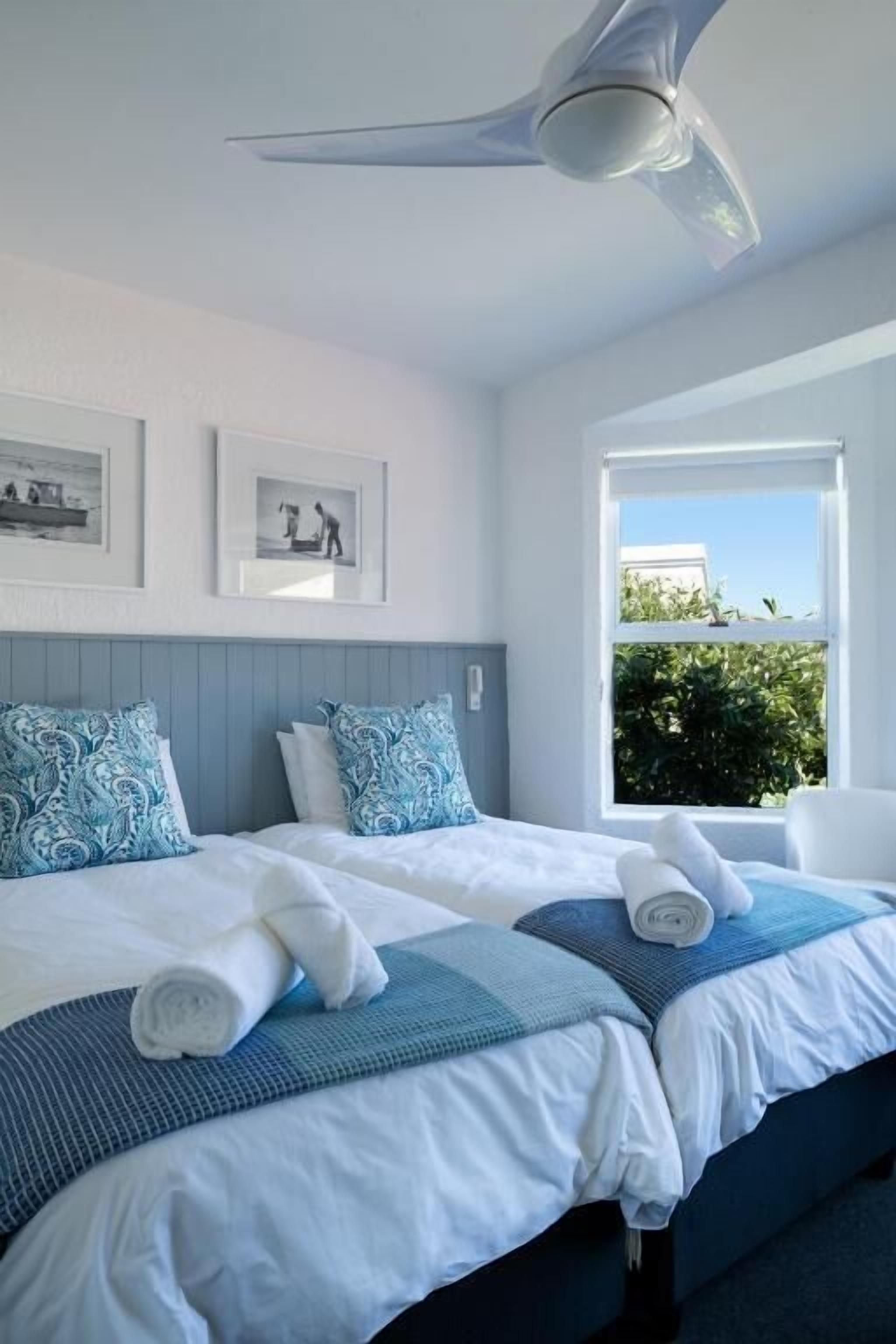 A hotel room with two beds, a ceiling fan, framed pictures on the wall, and a window with a view of greenery.