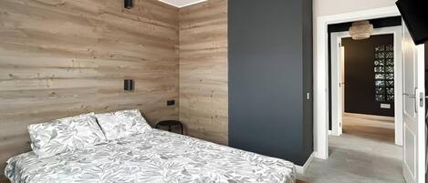 Una camera da letto con un letto, pannelli in legno alle pareti, una parete color grigio e una porta che dà su un'altra stanza.