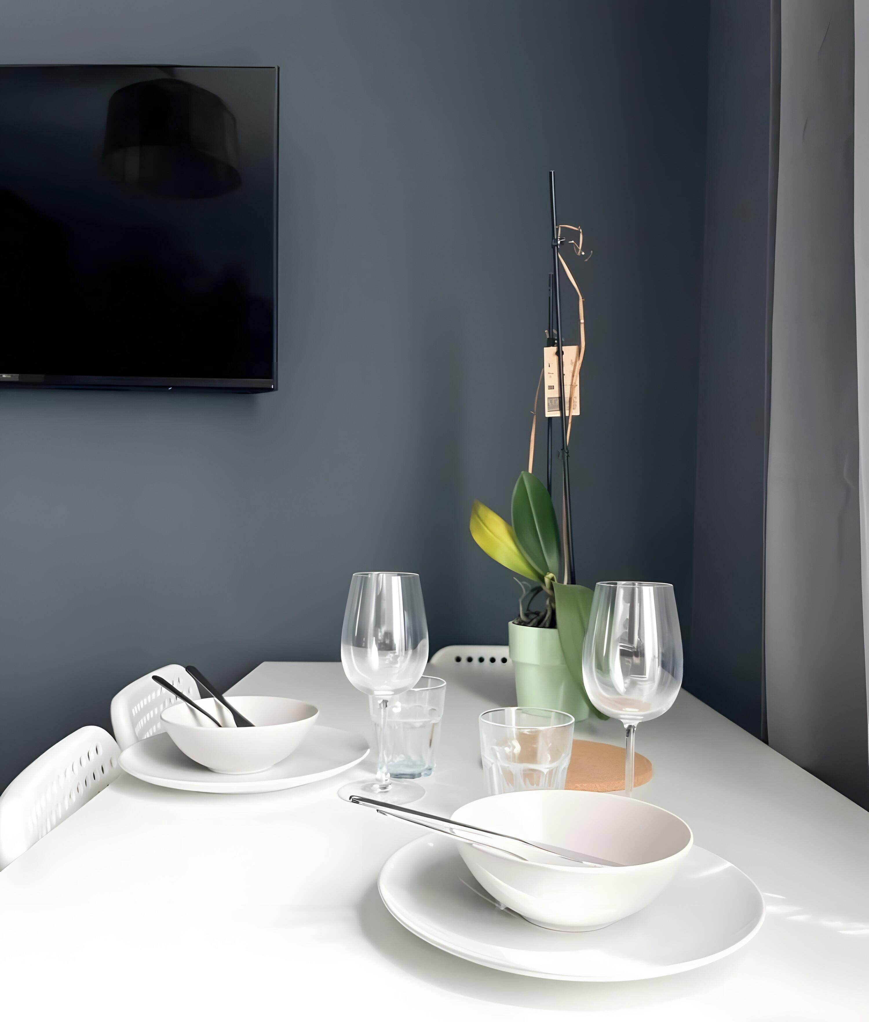 Un comedor moderno con una mesa dispuesta para dos personas, una planta en maceta y un televisor montado.