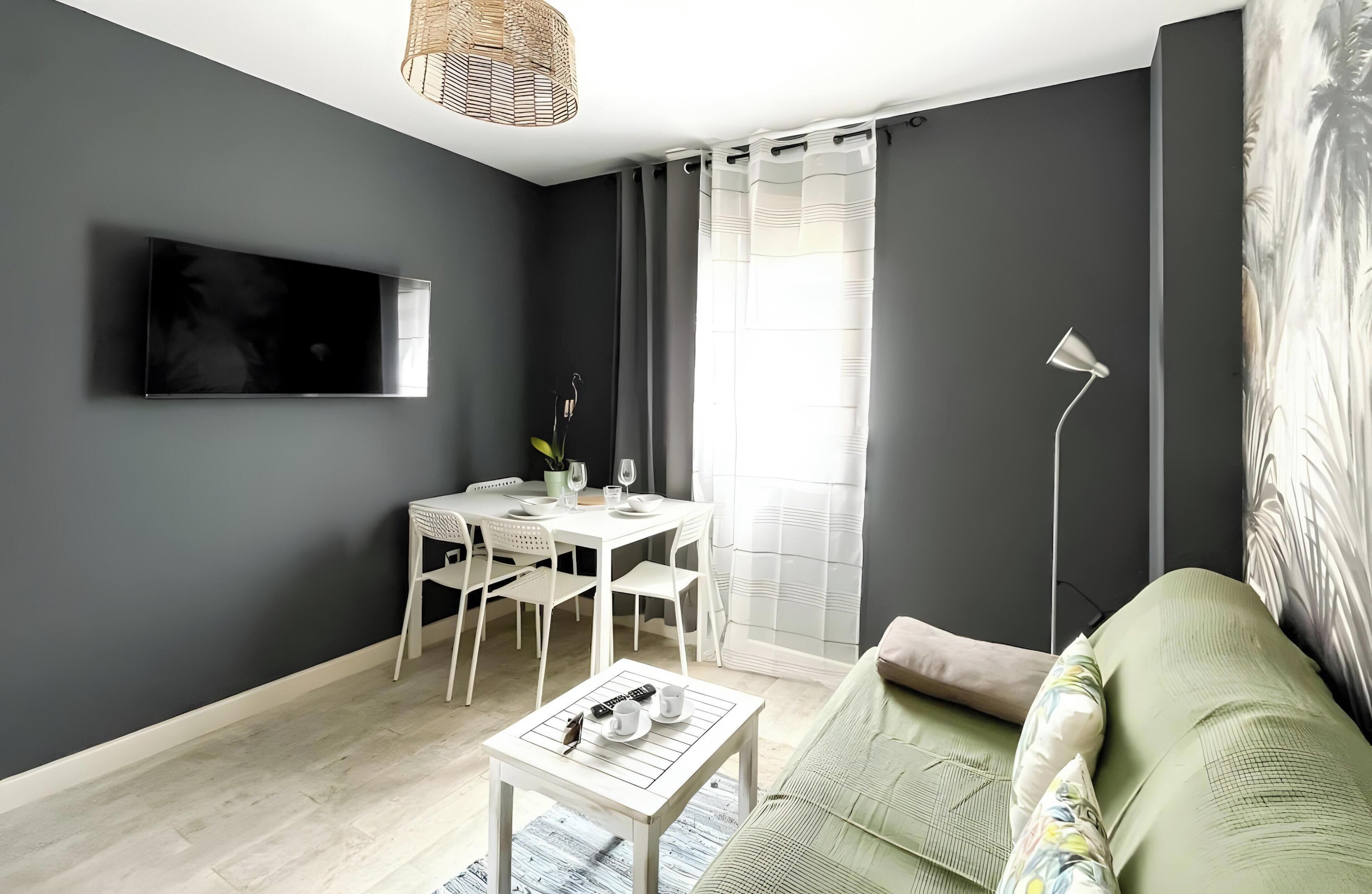 Una sala moderna con comedor, un televisor de pantalla plana montado en la pared, una lámpara de pie y un sofá con un cojín estampado.