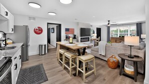 Una cocina moderna con horno empotrado, un comedor con mesa de madera y taburetes de bar, una sala de estar con sofá y televisor, y un ventanal con vistas.