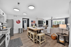 A modern kitchen with a built-in oven, a dining area with a wooden table and bar stools, a living room with a sofa and a TV, and a large window with a view. - Bama Oasis | 3 Private Suites | Pool & Fitness Center (Tuscaloosa)