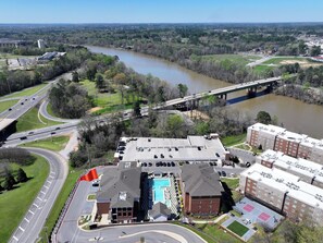 An aerial view of a building with a swimming pool, parking lot, and a river in the background. - Bama Oasis | 3 Private Suites | Pool & Fitness Center (Tuscaloosa)
