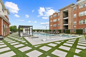 A pool area with a pergola, lounge chairs, and a brick building with balconies.