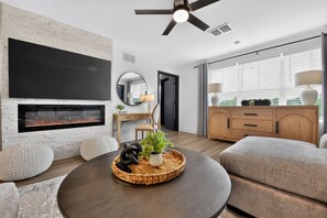 Uma sala de estar moderna com lareira, uma mesa de centro redonda com objetos decorativos, um sofá e uma televisão fixada na parede.