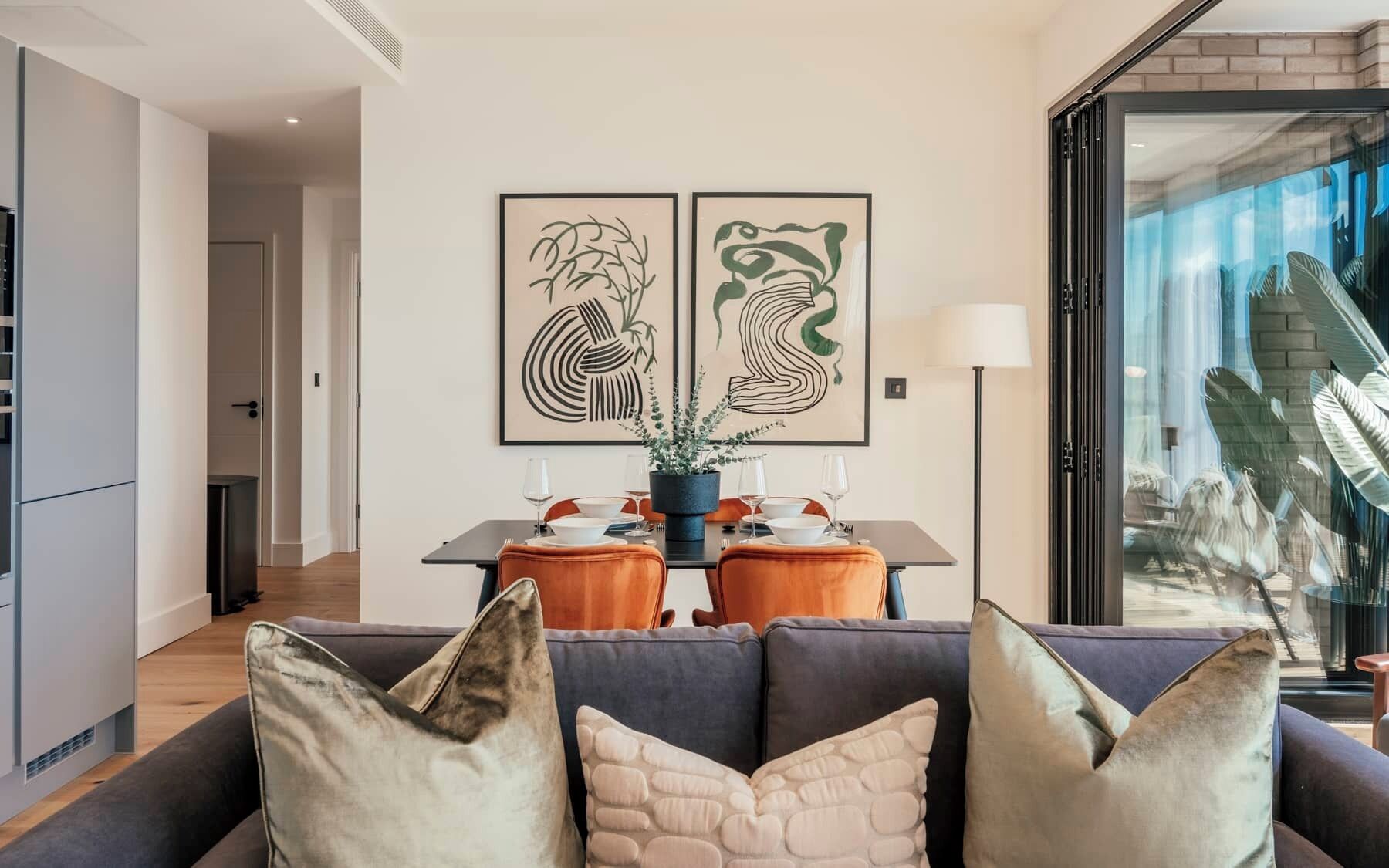 Un soggiorno moderno con zona pranzo, caratterizzato da un divano con cuscini decorativi, un tavolo da pranzo imbandito per quattro persone e due opere d'arte astratte appese alle pareti.