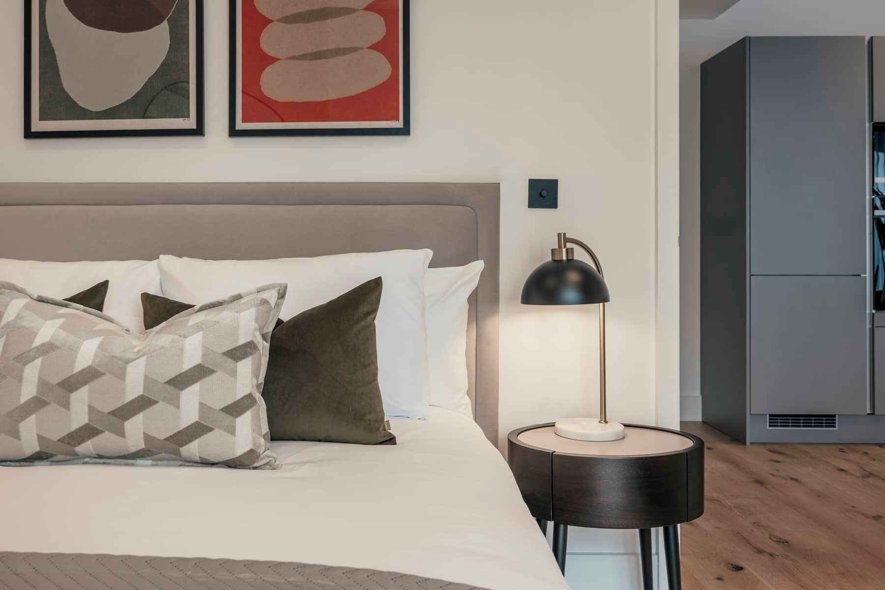 Un dormitorio moderno con cama, almohadas, una lámpara de noche y una obra de arte en la pared.
