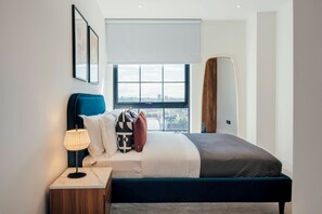A modern bedroom with a large window, a bed with multiple pillows, a bedside lamp, and a wooden nightstand. - Urban Rest Albert Embankment (London)