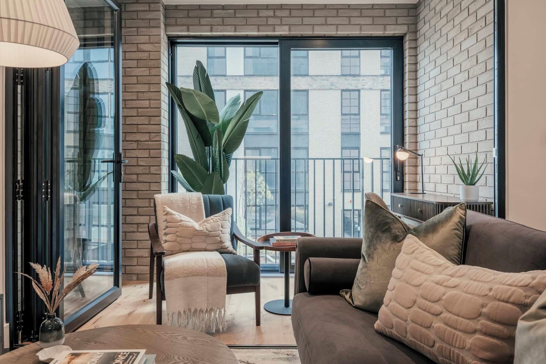 Un salon moderne avec un canapé, un fauteuil et une table basse. Une grande fenêtre offre une vue sur les immeubles et une plante en pot est posée à proximité.