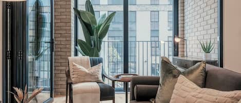 Ein modernes Wohnzimmer mit einer Couch, einem Sessel und einem Couchtisch. Es gibt ein großes Fenster mit Blick auf Gebäude und eine Küstpflanze.