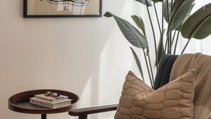 A room with a framed abstract art piece, a large potted plant, a round table with a book and glasses, and a cushioned chair with a throw blanket. - Urban Rest Albert Embankment (London)