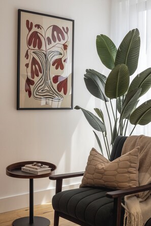 Ein Zimmer mit einem gerahmten abstrakten Kunstwerk, einer großen Kübelpflanze, einem runden Tisch mit einem Buch und Gläsern sowie einem gepolsterten Sessel mit einer Decke.