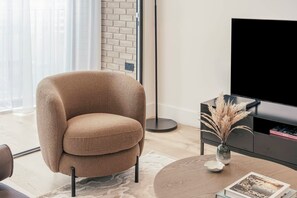 Ein modernes Wohnzimmer mit einem beigen Sessel, einem runden Couchtisch, einer schwarzen TV-Konsole und einer Vase mit Trockenblumen.