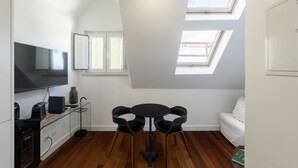 A modern kitchen with a flat-screen TV, a coffee machine, and a glass cabinet. There is a dining area with a round table and chairs, and a white sofa. The room has skylights and a view of the outside. - Estrela Riverside Studios IV by Homing (Lisbon)