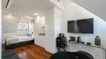 Quarto moderno com cama, escrivaninha com computador, televisão e janela com vista para o telhado.
