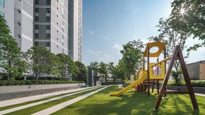 A playground with a yellow slide and swings, surrounded by a well-maintained lawn and trees, with a tall building in the background. - Pattaya Central I Near The Beach I Nice Pool (Pattaya)