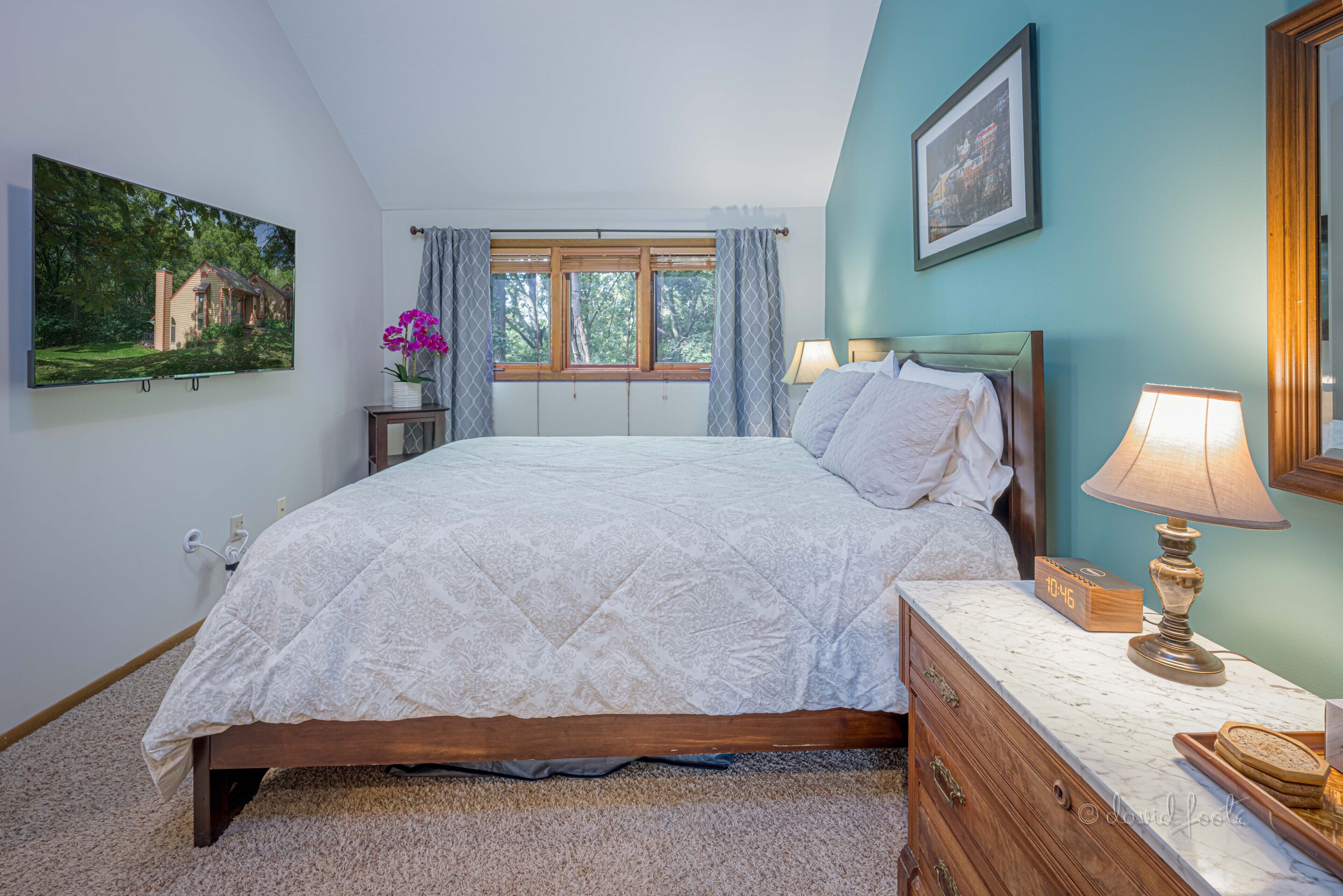 A bedroom with a bed, bedside tables, a nightstand, a lamp, a clock, and a framed picture on the wall.