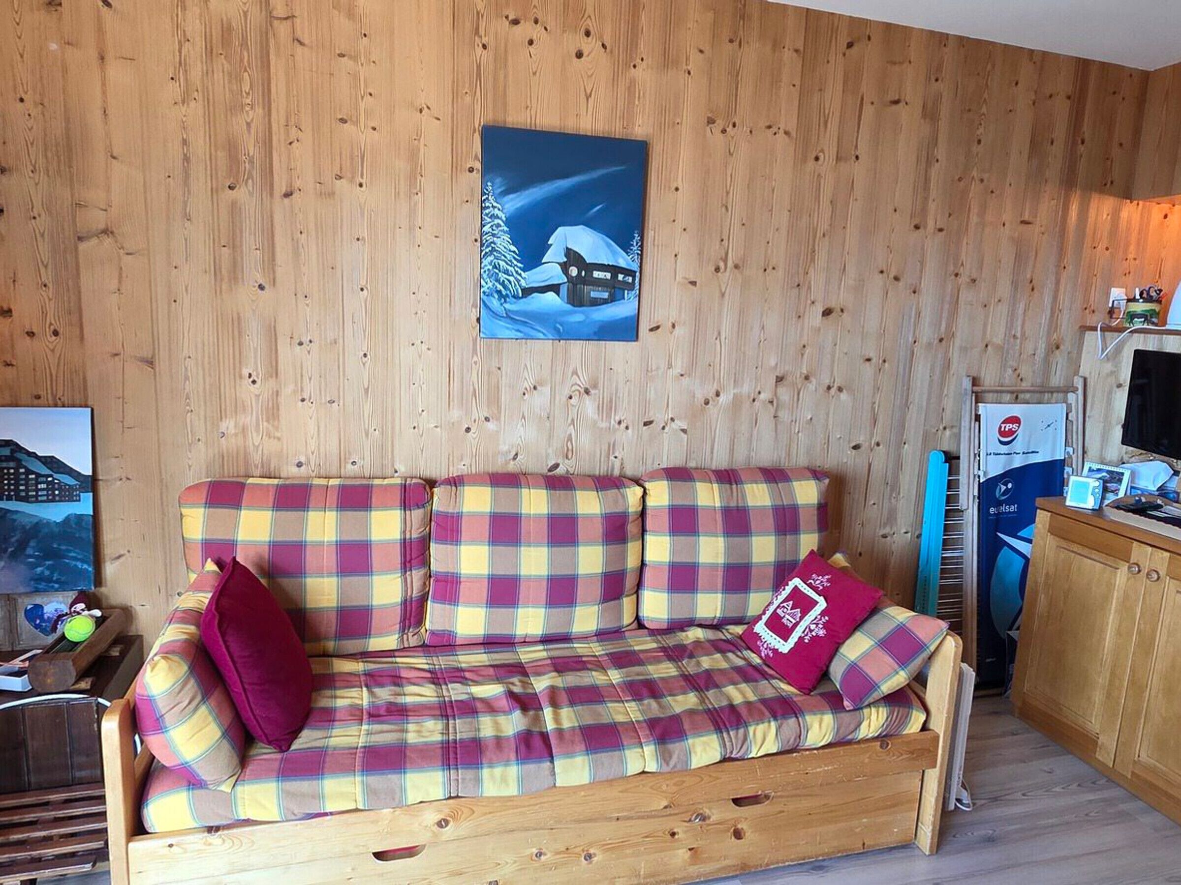 Una habitación con paneles de madera, un sofá de cuadros, un cuadro de un paisaje nevado y una pequeña cocina.