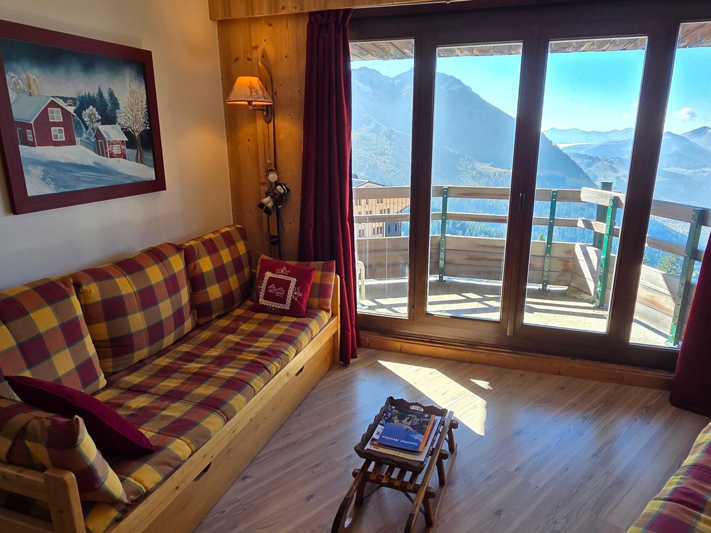 Une pièce chaleureuse avec un plancher en bois, un canapé à carreaux, une petite table avec un livre, un tableau représentant un paysage enneigé et une vue sur les montagnes par de grandes fenêtres.