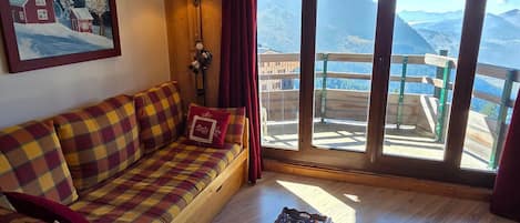 Un'accogliente stanza con pavimento in legno, un divano a quadri, un tavolino con un libro, un quadro raffigurante una scena innevata e vista sulle montagne attraverso ampie finestre.