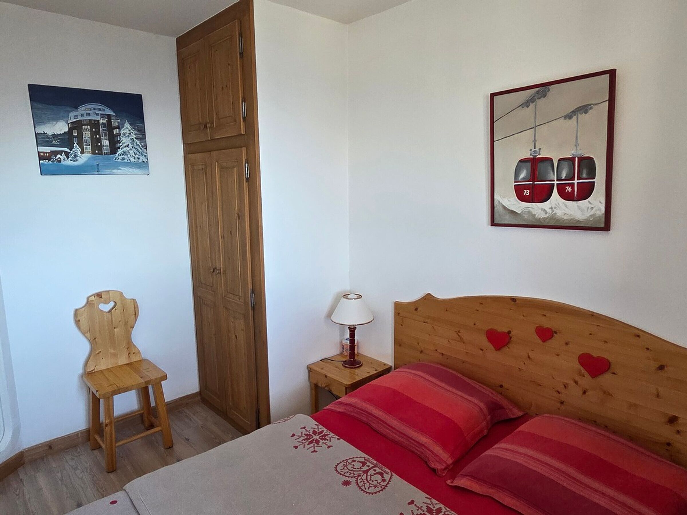 Une chambre à coucher comprenant un lit, une chaise en bois, une table de chevet, une lampe et deux œuvres d’art accrochées au mur.