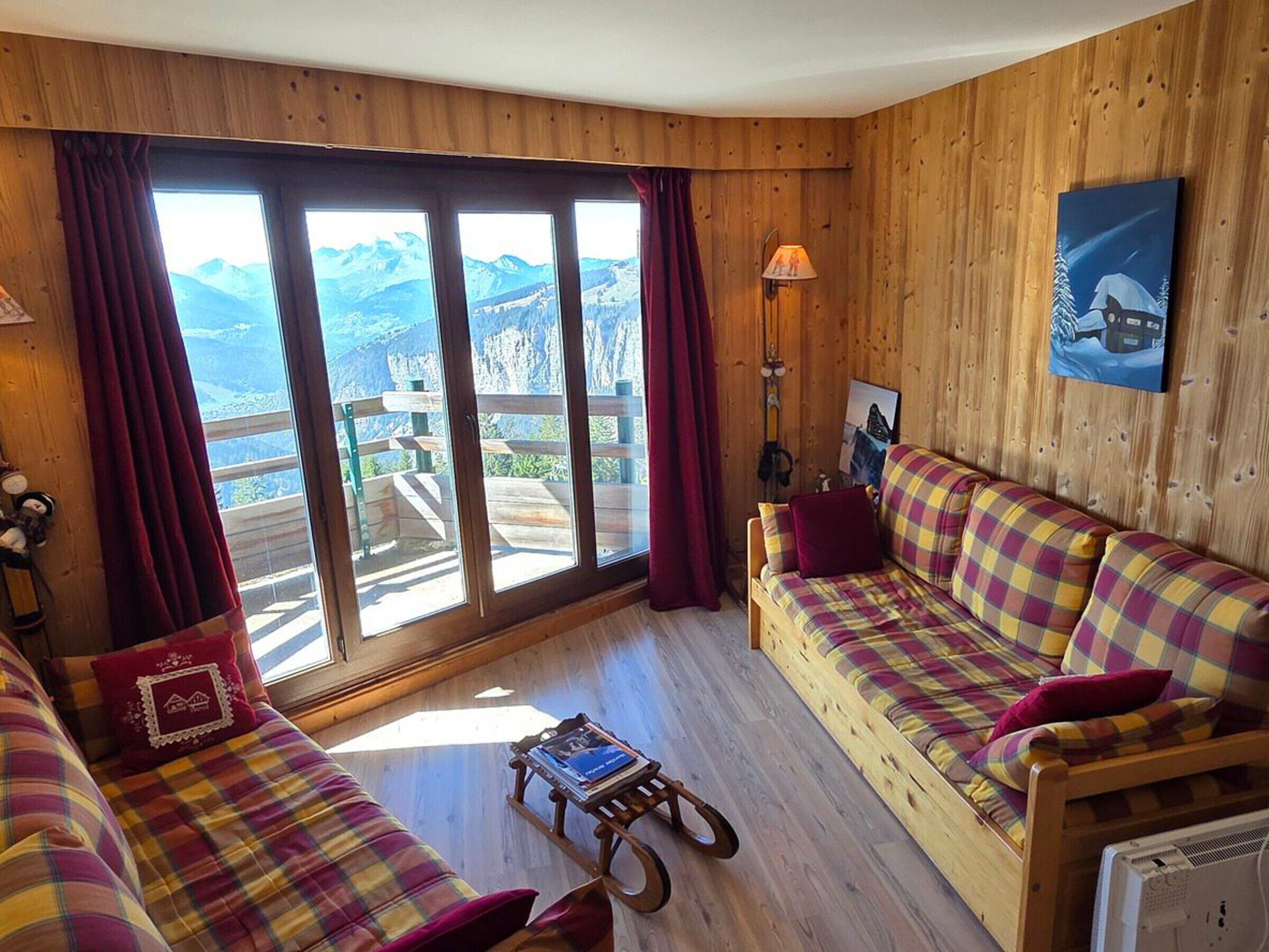 Quarto aconchegante com decoração em madeira, um sofá, uma pequena mesa e vista para montanhas através de grandes janelas.