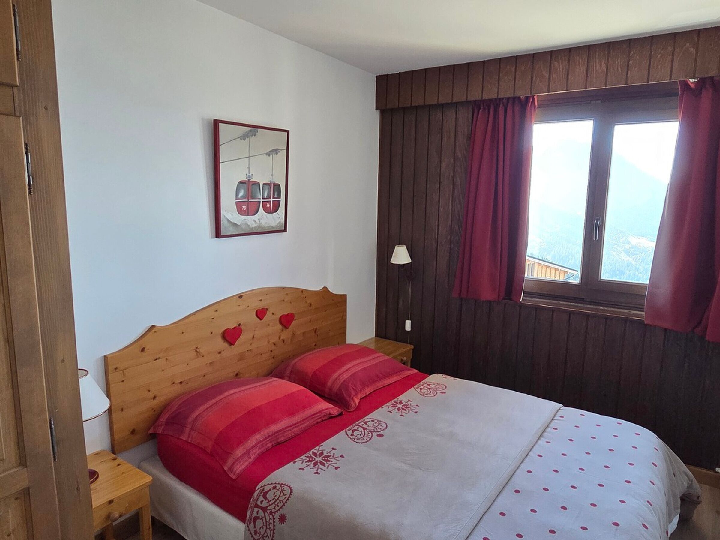 Quarto com cabeceira de madeira, cama com roupa de cama vermelha e branca, mesa de cabeceira, abajur, janela com cortinas e um quadro pendurado na parede.